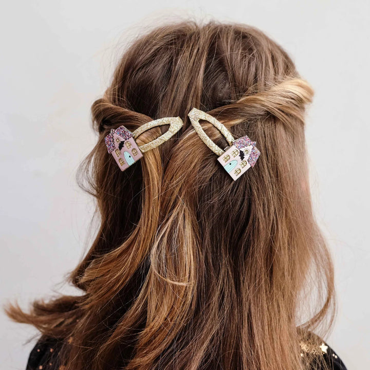 A close-up of a person's brown hair styled with two decorative hair clips. The clips have glittery gold arches and rectangular pastel-colored charms featuring haunted houses shown against a plain light gray background.
