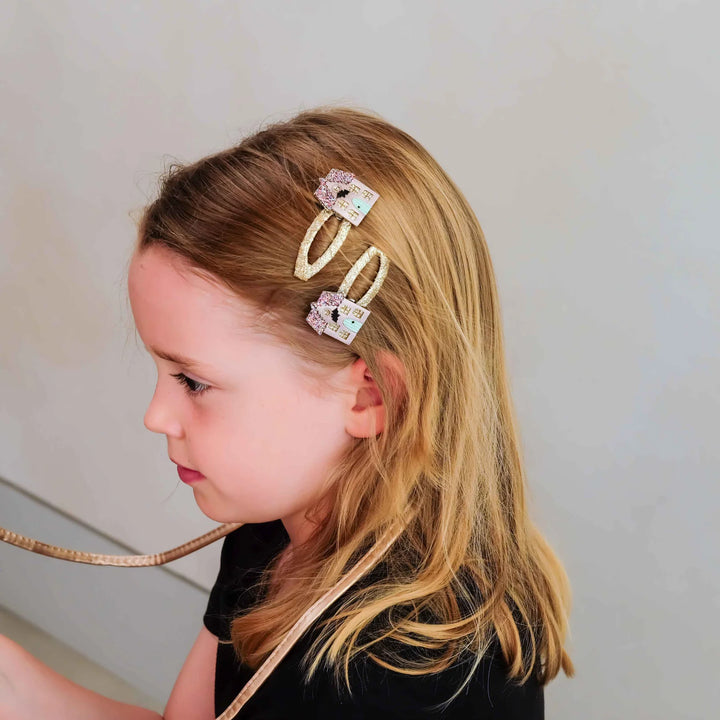 A young girl with blonde hair wearing three decorative hair clips, shown in profile against a light gray background.