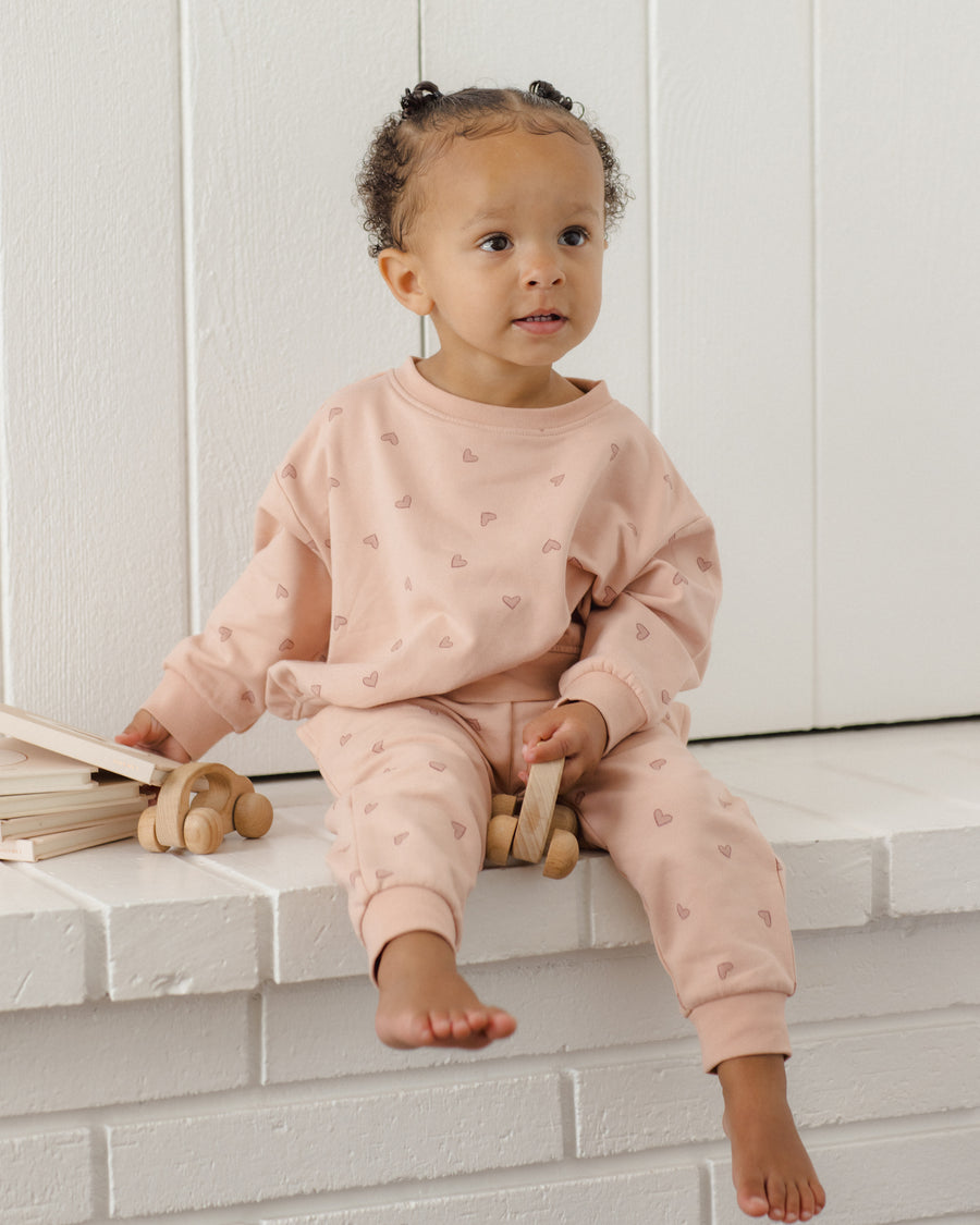A toddler girl sits barefoot on a white brick ledge wearing a light pink long-sleeve sweatshirt and matching jogger pants with a small heart pattern. She is holding a wooden toy, with a stack of wooden toys on the left, against a white paneled wall background.