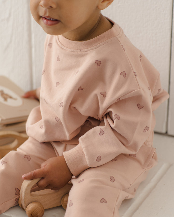 A baby wearing a light pink long-sleeve sweatshirt and matching pants set, both decorated with a small repeating heart pattern. The baby is sitting on a white surface next to a wooden toy car.