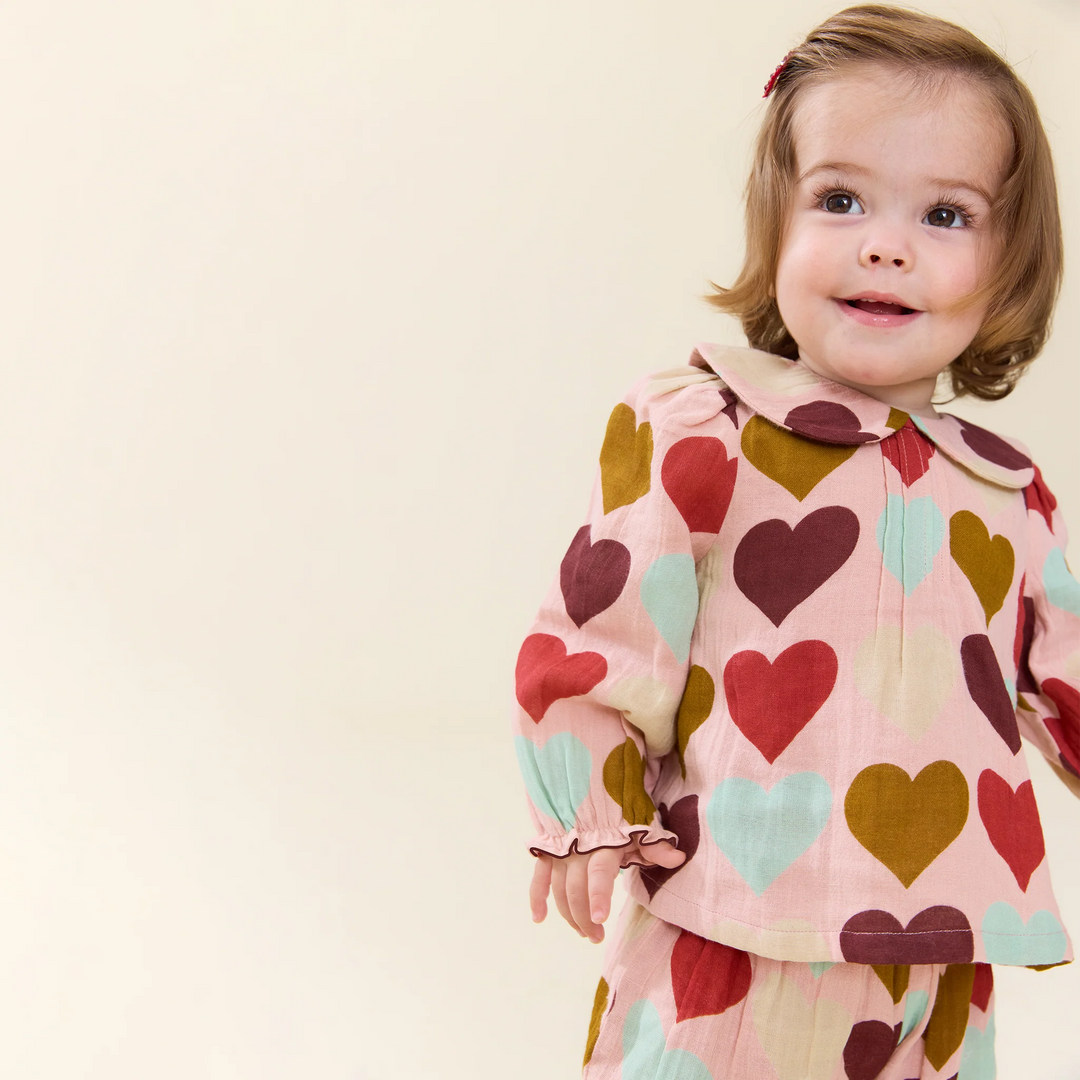 A toddler girl wearing a matching long-sleeve top and skirt set with a pink base and a heart pattern in red, mustard, mint, and burgundy, posed against a solid cream background.