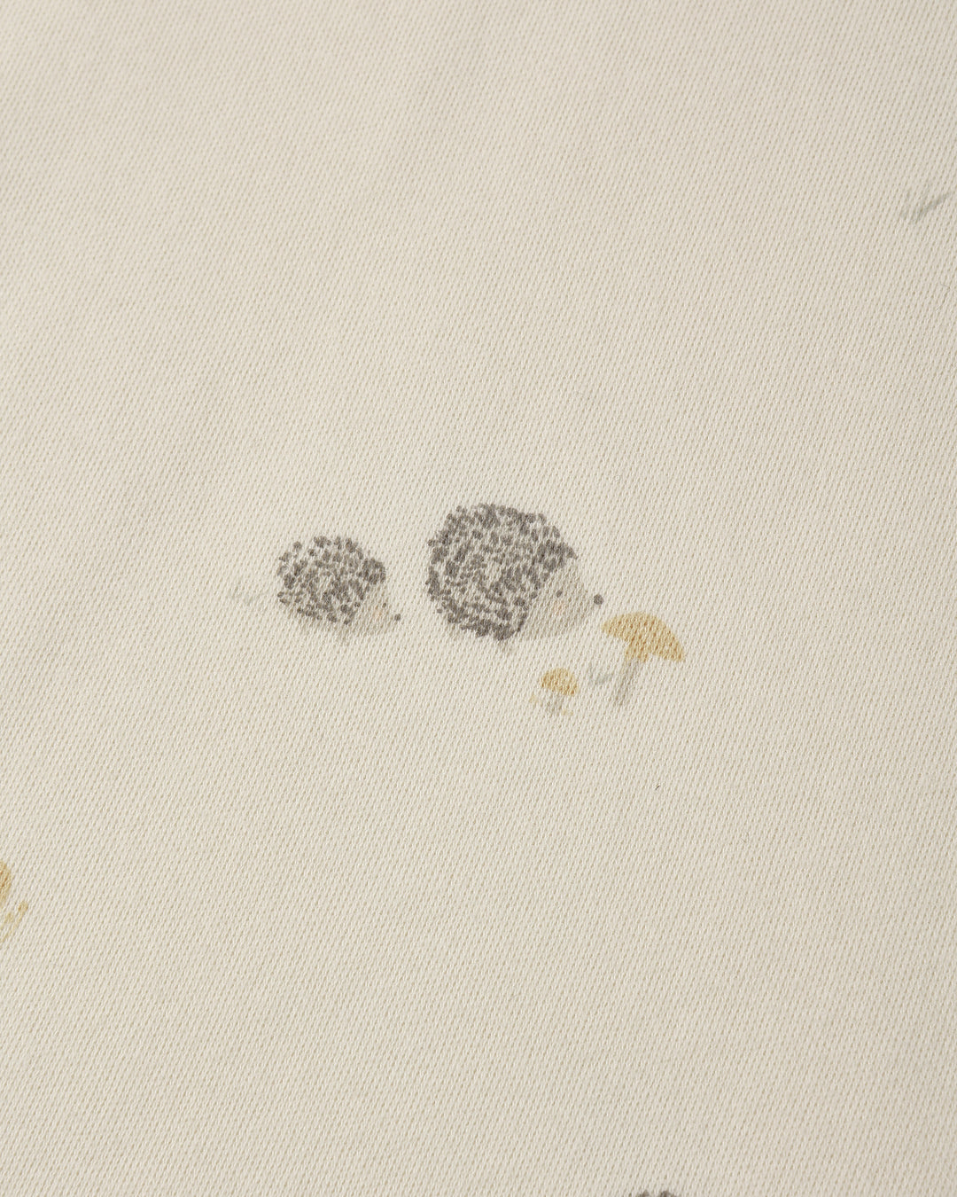Close-up of a light cream fabric with a small printed illustration of two hedgehogs and a mushroom near the center.