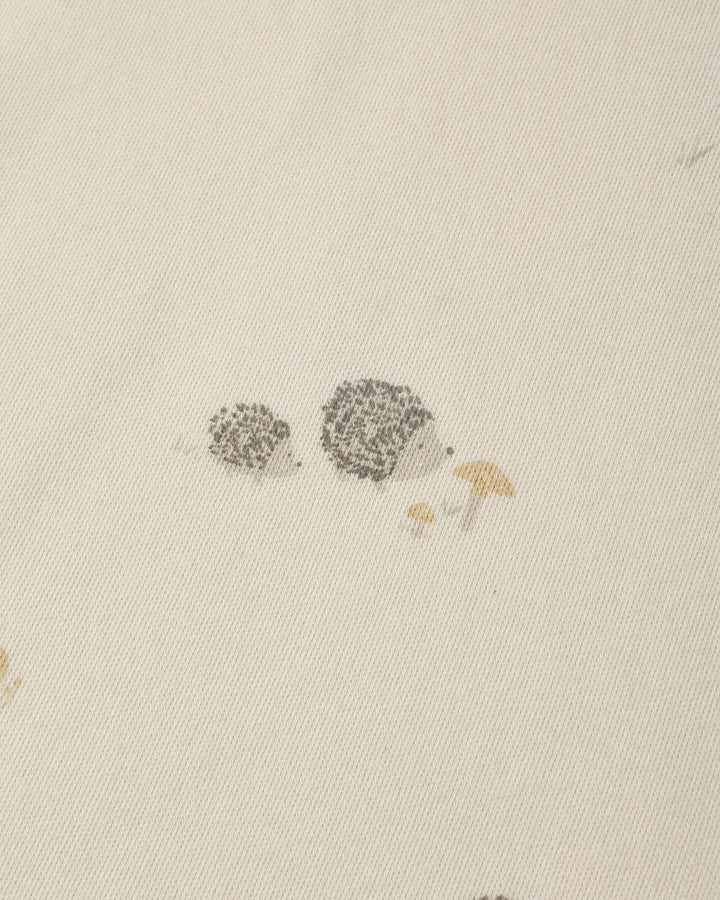 Close-up of a light cream fabric with a small printed illustration of two hedgehogs and a mushroom near the center.