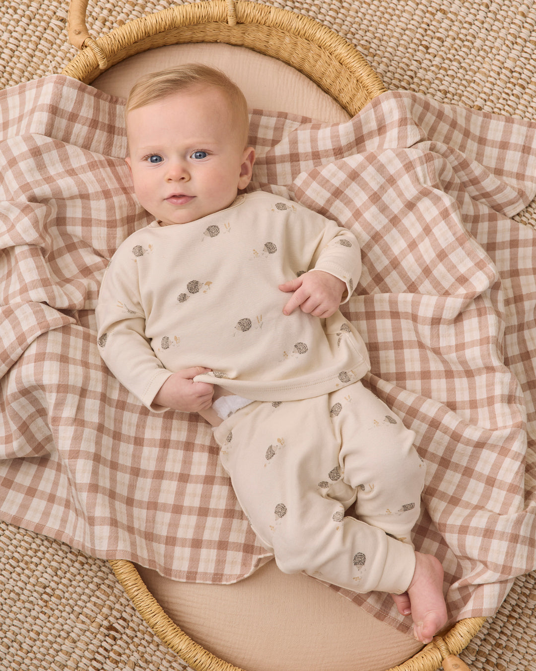 A baby lying on a woven mat under a beige and brown gingham blanket, wearing a cream long-sleeve pajama set with small gray animal prints.