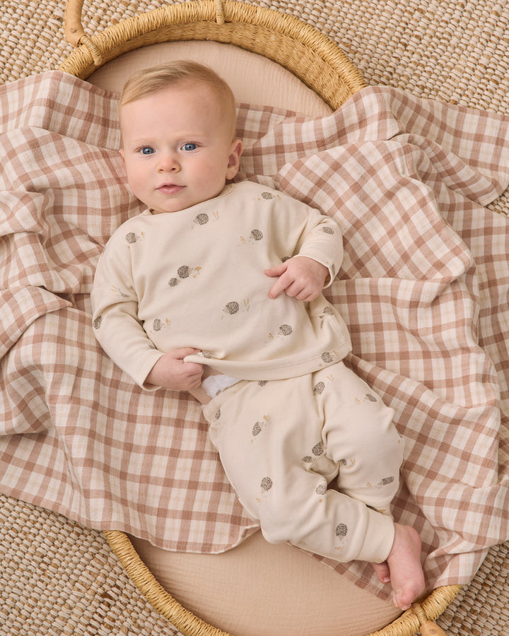 A baby lying on a woven mat under a beige and brown gingham blanket, wearing a cream long-sleeve pajama set with small gray animal prints.