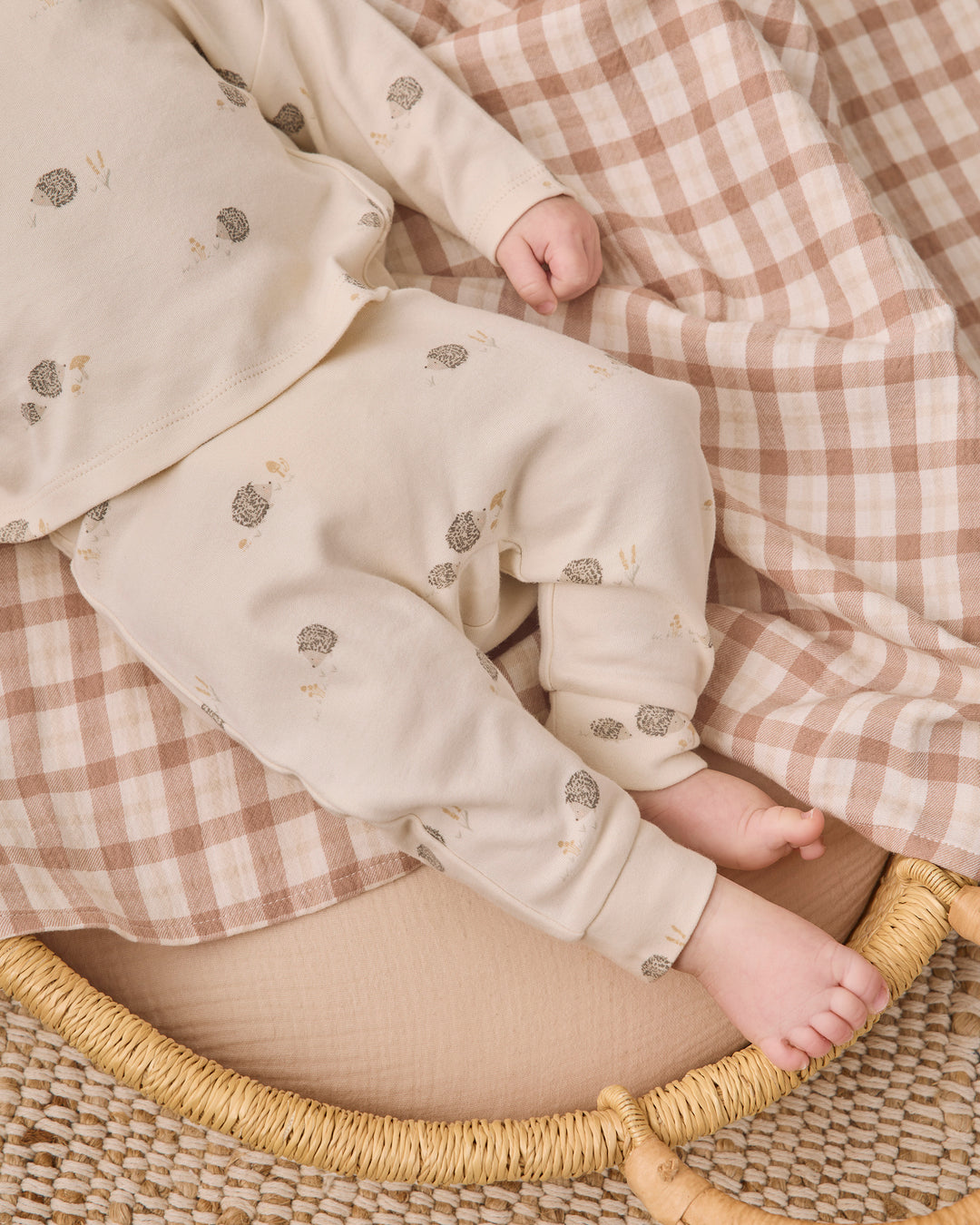 A baby wearing a cream-colored long-sleeve romper and matching pants with small gray and beige floral prints, lying on a brown and white checkered blanket inside a woven rattan bassinet.