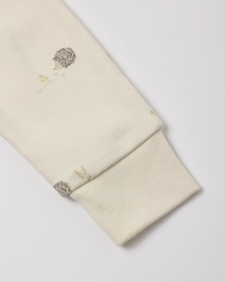 Close-up of a cream-colored knit sleeve with a ribbed cuff, featuring a small embroidered hedgehog design in gray and yellow.