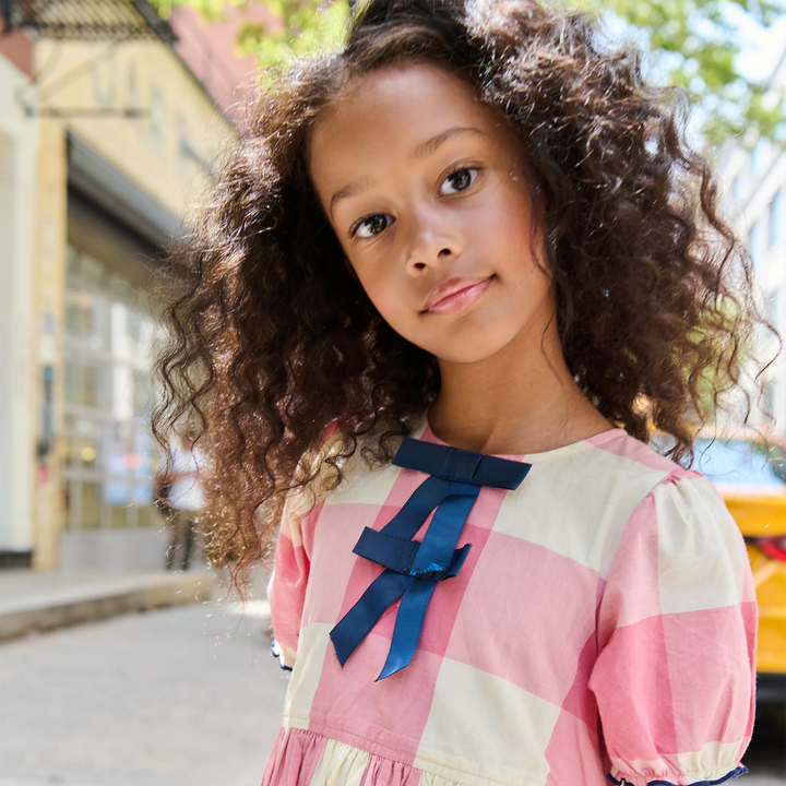 A young girl with voluminous curly brown hair wearing a pink and cream gingham dress with a navy blue bow on the chest, photographed outdoors on a sunny street.