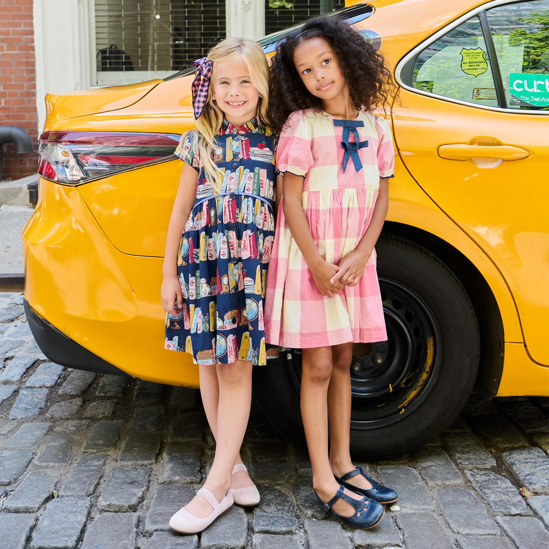 Two young girls standing in front of a bright yellow car on a cobblestone street, wearing matching patterned dresses. The girl on the left wears a navy dress with a bookshelf print and a purple bow, and the girl on the right wears a pink and white gingham dress with a blue bow.