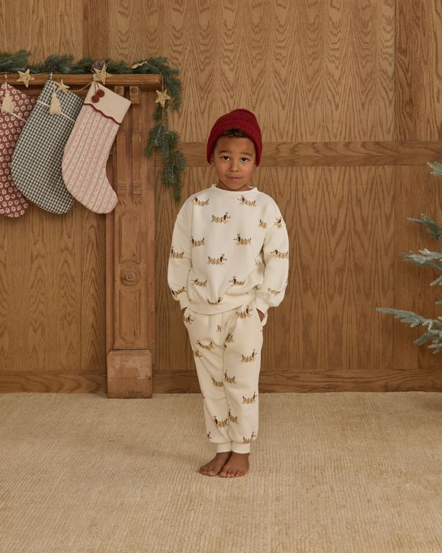 Child wearing a christmas dog  patterned outfit standing in front of wooden paneling with stockings.