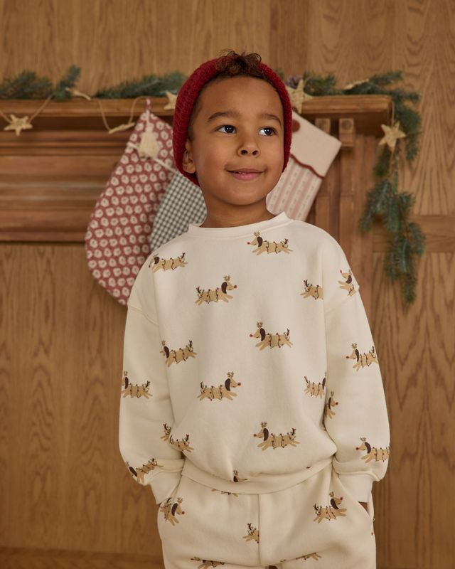 Child wearing a cream-colored outfit with christmas dog pattern in front of a wooden wall with Christmas decorations.