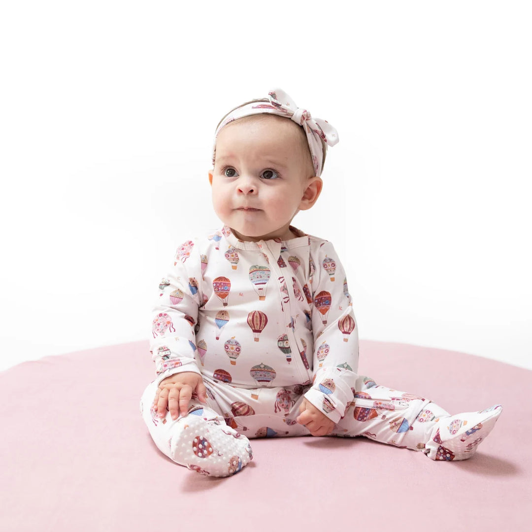 A baby sitting on a pink circular mat wearing a cream-colored footed sleeper with a colorful hot air balloon pattern and a matching bow headband.