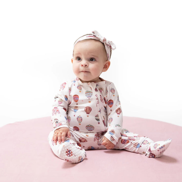A baby sitting on a pink circular mat wearing a cream-colored footed sleeper with a colorful hot air balloon pattern and a matching bow headband.