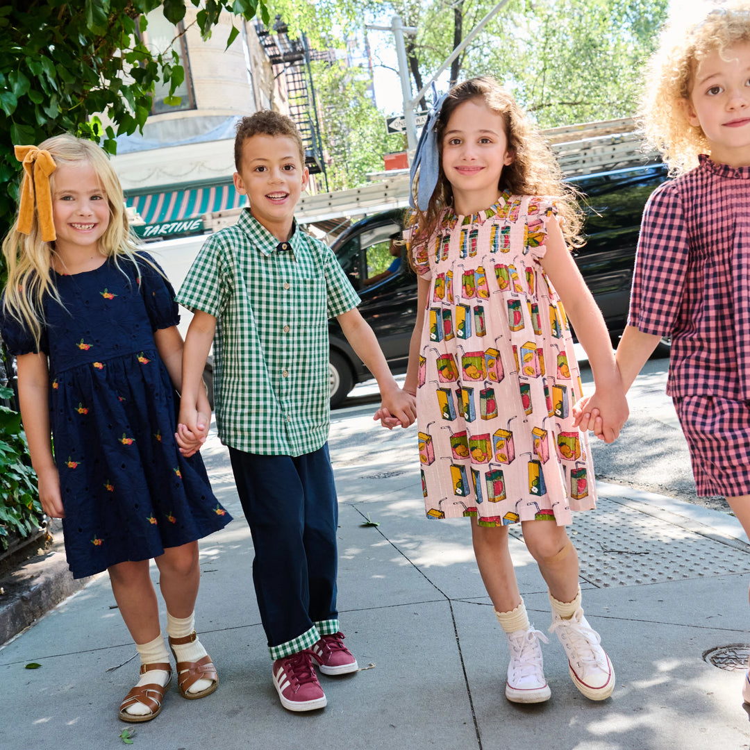 Four children holding hands walking hand-in-hand on a sunny city sidewalk. They are wearing matching but distinct outfits: a navy dress with yellow accents, a green gingham shirt and navy pants, a pink dress with a colorful retro pattern, and a red gingham shirt and pink shorts.