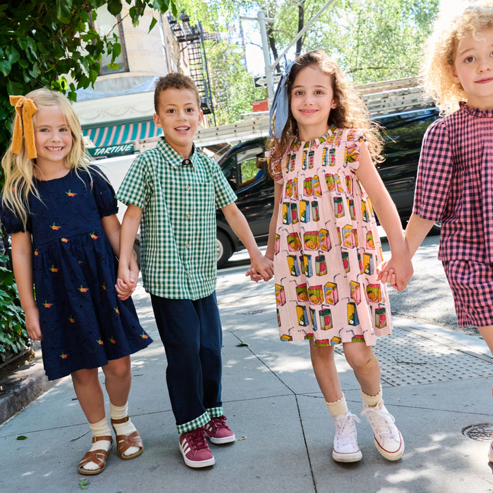 Four children holding hands walking hand-in-hand on a sunny city sidewalk. They are wearing matching but distinct outfits: a navy dress with yellow accents, a green gingham shirt and navy pants, a pink dress with a colorful retro pattern, and a red gingham shirt and pink shorts.
