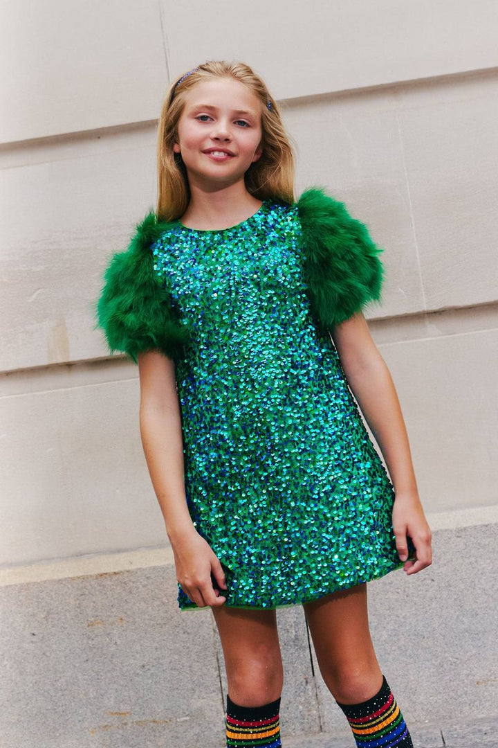 A young girl modeling a green sequined dress with fluffy feathered green sleeves, standing against a beige stone wall.