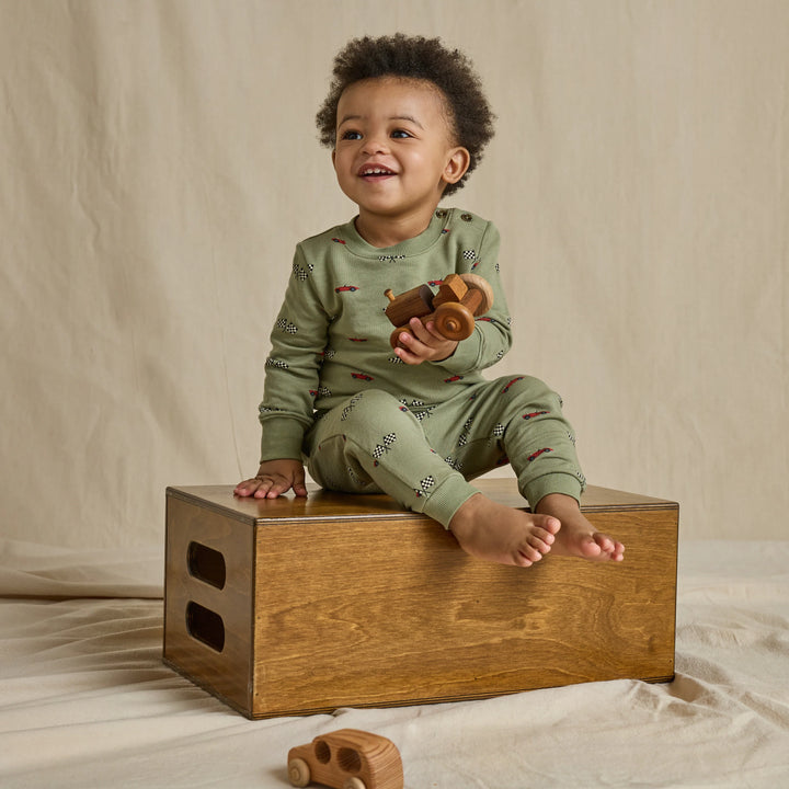 A smiling toddler sits on a wooden box wearing an olive green long-sleeve pajama set with a small red and black pattern. The child holds a wooden toy car in a studio setting with a beige fabric backdrop.