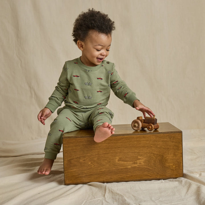 A toddler wearing sage green long-sleeve pajamas with a small red and black pattern sits on a wooden box, reaching for a wooden toy car in a studio setting with a beige fabric backdrop.