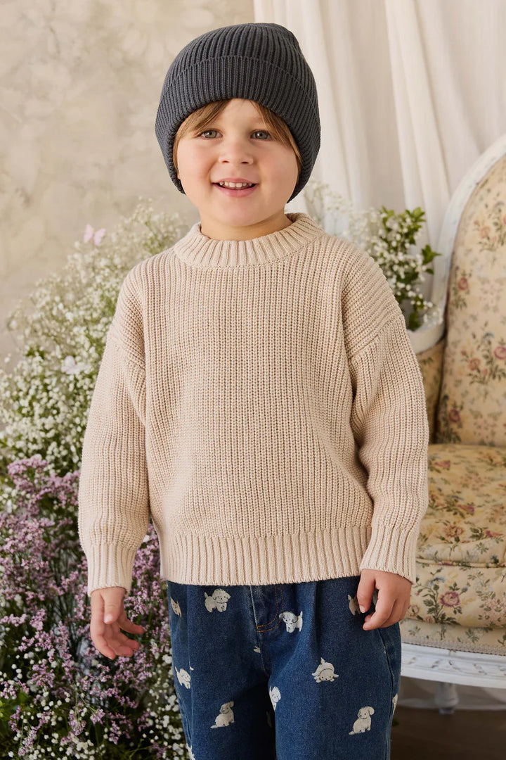 Child wearing a beige sweater and dark beanie standing in a decorative room with flowers and a sofa.