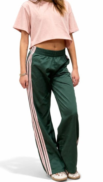 A model wearing dark green wide-leg track pants with pink and white side stripes, paired with a light pink cropped t-shirt and white sneakers, shown against a plain white background.
