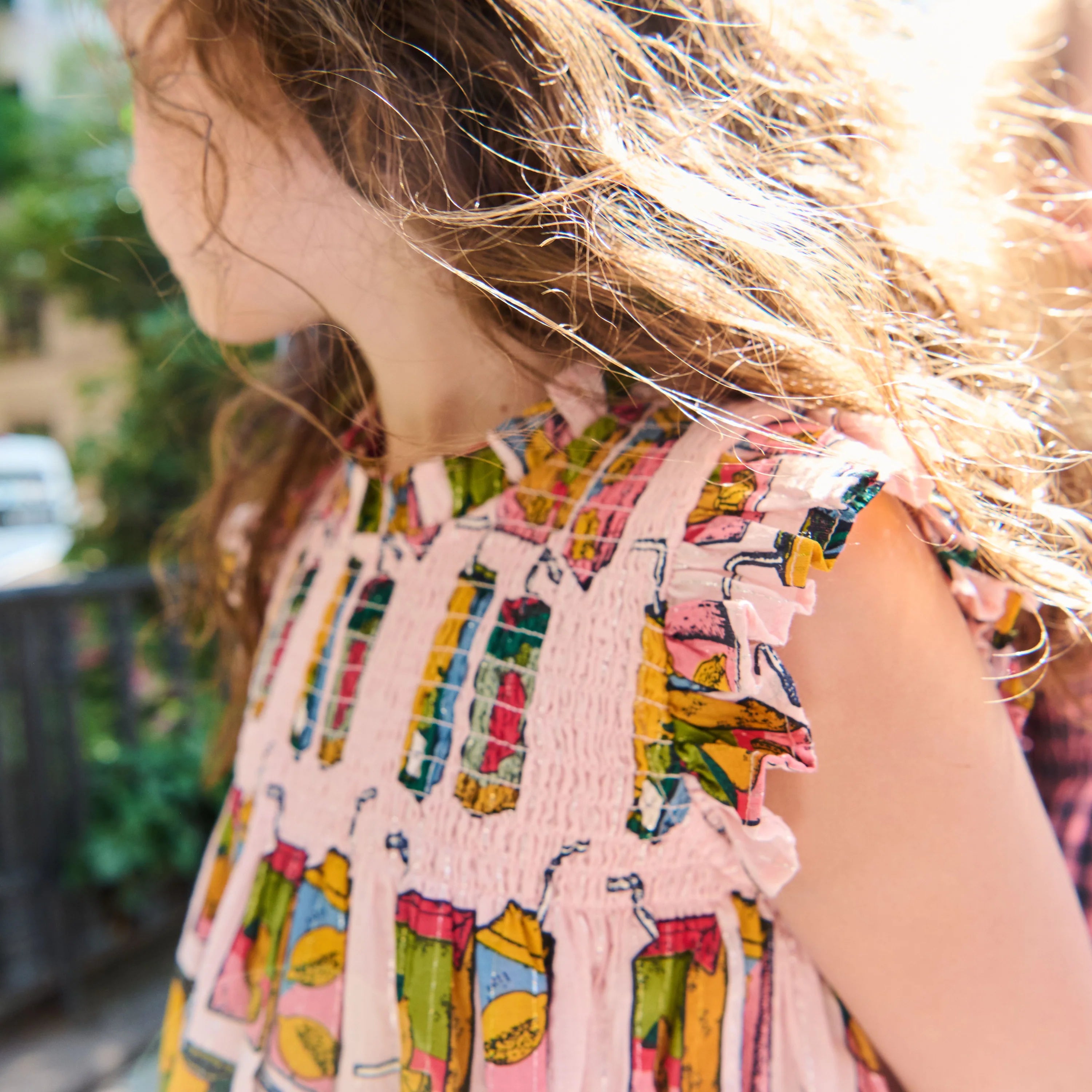 A close-up side view of a child wearing a light pink sleeveless top with a colorful illustrated pattern of drinks and ruffled shoulder details, outdoors with bright sunlight and blurred greenery in the background.