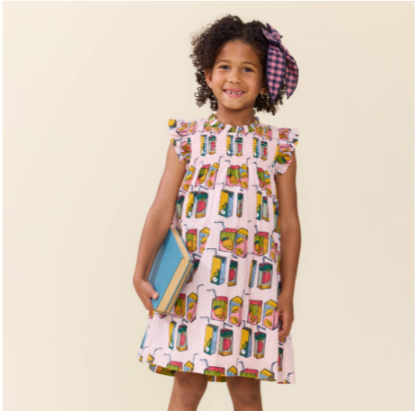 A young girl with curly hair wearing a pink sleeveless dress with a colorful juice box print and a purple plaid bow headband, holding a blue book against a solid beige background.