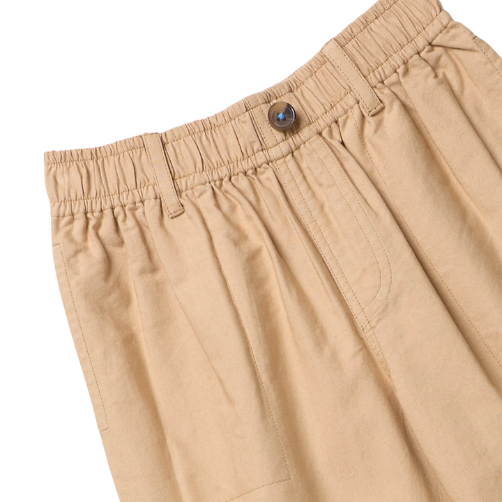 Close-up of beige pleated trousers showing the elastic waistband, button closure, and side pocket on a white background.