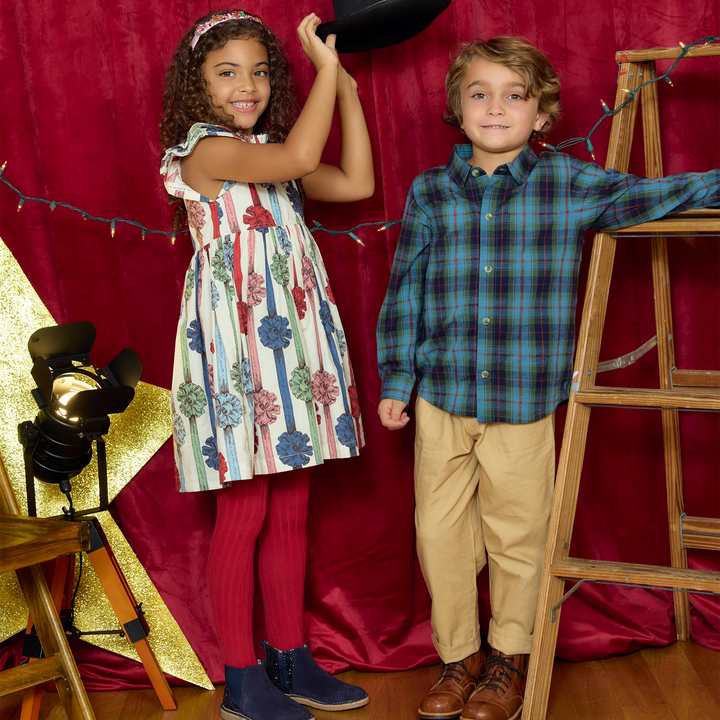 A young girl in a floral sleeveless dress and red tights holds a black cowboy hat next to a young boy in a blue-green plaid button-down shirt and khaki pants, posed in front of a red curtain backdrop with a studio light and wooden ladder.