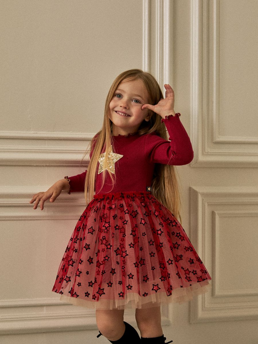 A young girl with long blonde hair wearing a festive red dress with a star-patterned tulle skirt and a gold star applique, posing against a beige paneled wall.