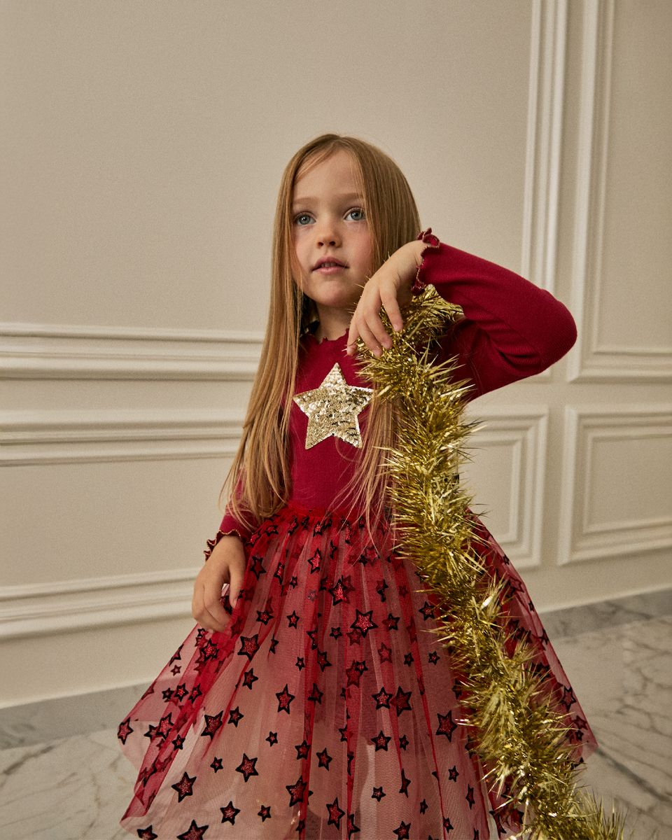 A young girl with long light brown hair stands indoors wearing a festive red dress with a star pattern and holding a gold tinsel garland. The dress features a solid red long-sleeve bodice with a large gold star applique at the chest, and a sheer red tulle skirt decorated with black star motifs.