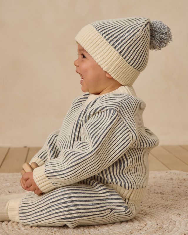 Baby wearing a striped sweater and hat sitting on a wooden floor.