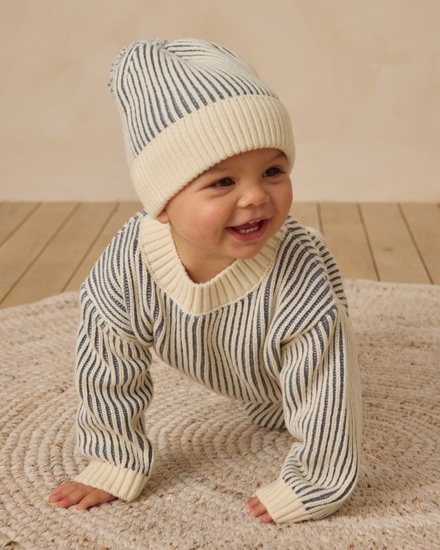 Baby wearing a striped sweater and hat on a wooden floor.