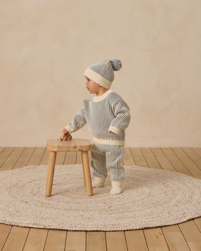 Child in striped outfit and knit hat standing on a wooden floor with a beige wall background