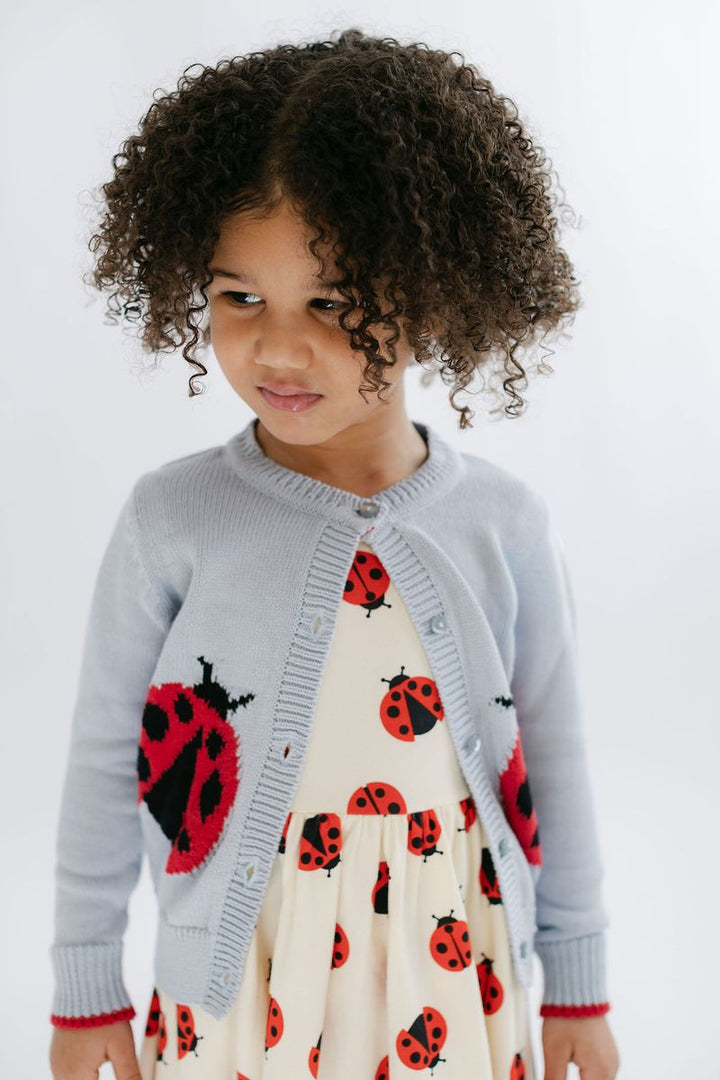 A young child with curly brown hair wearing a light blue cardigan over a cream dress with a red and black ladybug print, photographed against a plain light background.