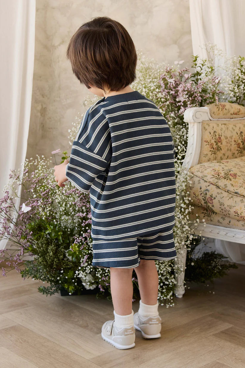Child in a striped outfit standing in a room with flowers and a chair.