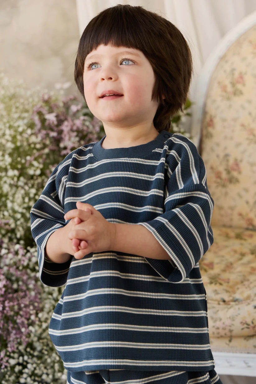 Child wearing a navy and white striped shirt standing in a decorative room with flowers and a chair.