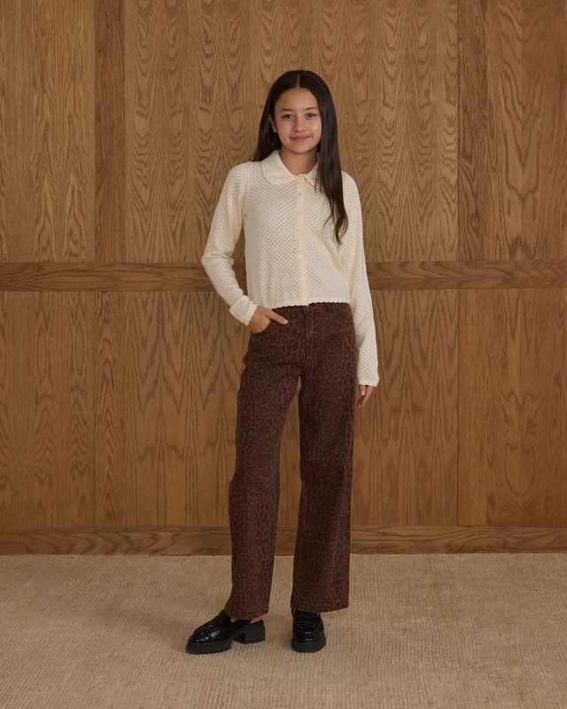 Person wearing a white blouse and brown patterned pants standing against a wooden paneled wall.