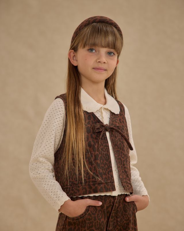 Young girl wearing a brown leopard vest with a patterned collar against a beige background