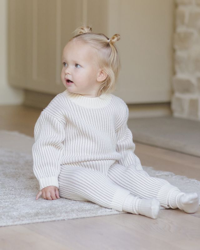 Child wearing a lilac contrast ribbed sweater and pants sitting on a rug indoors.