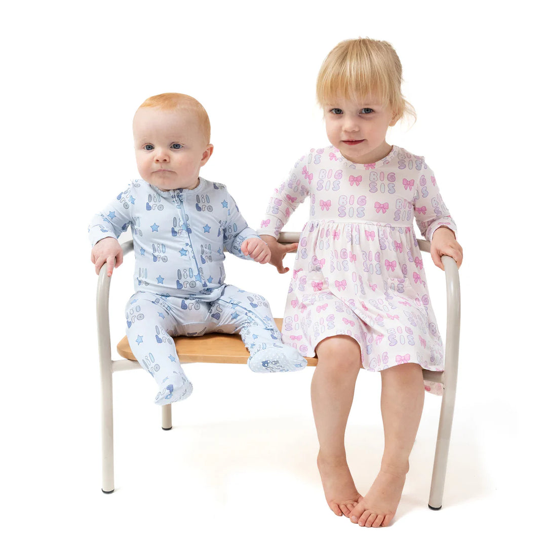 Two children sitting on a chair, one in blue pajamas and the other in a pink dress that says 'big sis' pattern.