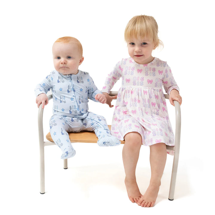 Two children sitting on a chair, one in blue pajamas and the other in a pink dress that says 'big sis' pattern.