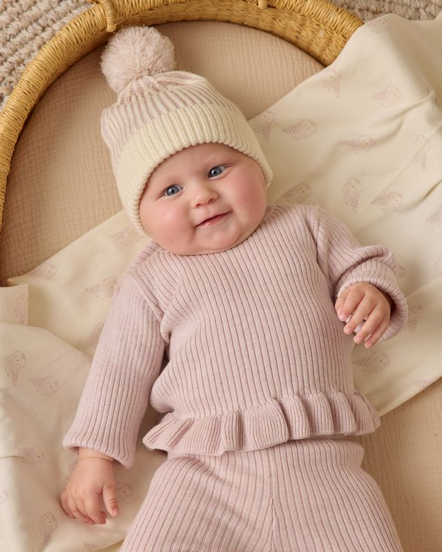 Baby in pink outfit and white knit hat with pom-pom in a wicker basket.