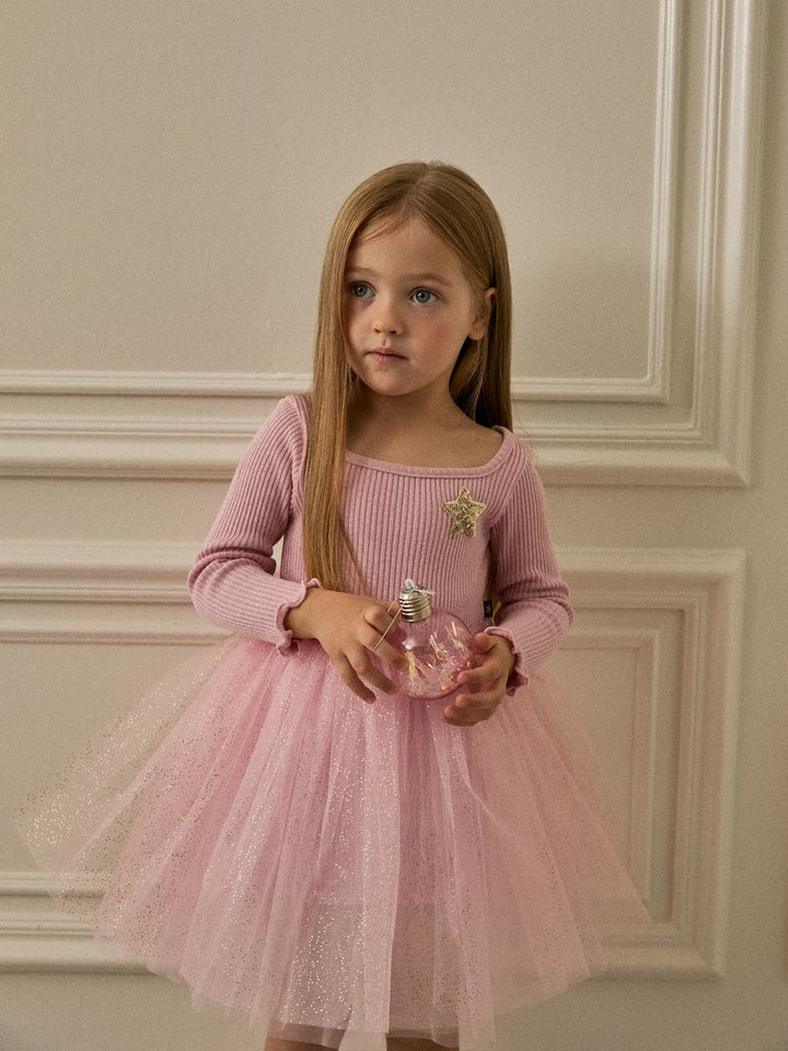 A young girl with long light brown hair stands against an off-white paneled wall, wearing a pink ribbed long-sleeve top and a matching pink tulle tutu skirt. She is holding a small pink perfume bottle.
