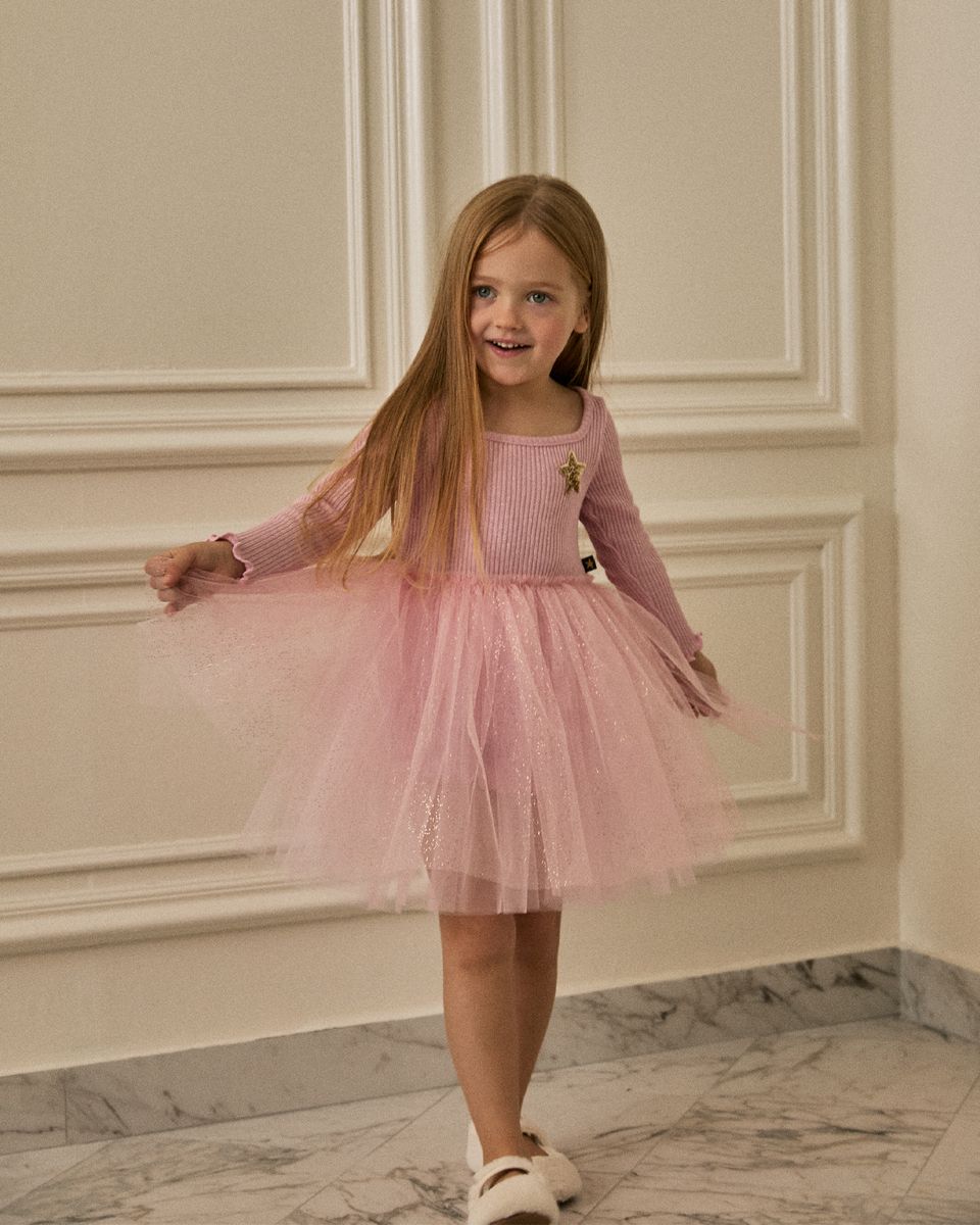 A young girl with long light brown hair stands indoors wearing a pink long-sleeve knit top and a matching pink tulle tutu skirt. She is smiling and holding out both sides of the skirt.