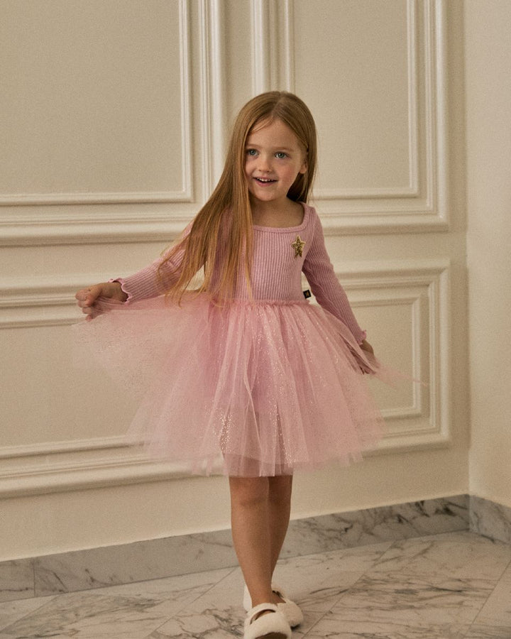 A young girl with long light brown hair stands indoors wearing a pink long-sleeve knit top and a matching pink tulle tutu skirt. She is smiling and holding out both sides of the skirt.