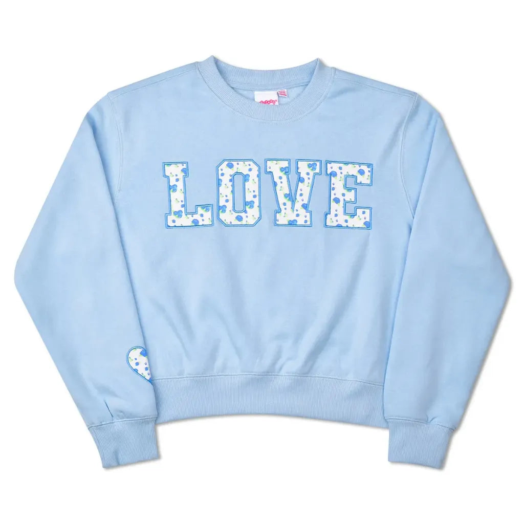 A light blue crewneck sweatshirt featuring the word 'LOVE' printed across the chest in a white floral pattern. A small matching floral heart patch is attached to the left sleeve.