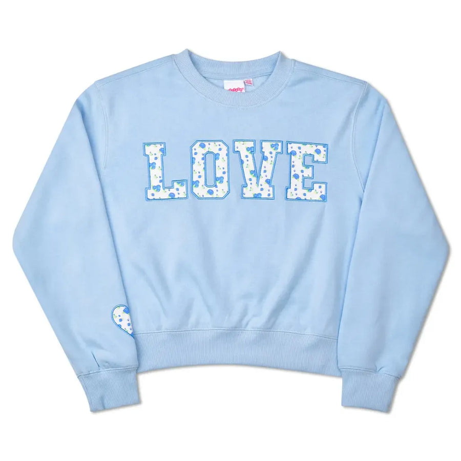 A light blue crewneck sweatshirt featuring the word 'LOVE' printed across the chest in a white floral pattern. A small matching floral heart patch is attached to the left sleeve.