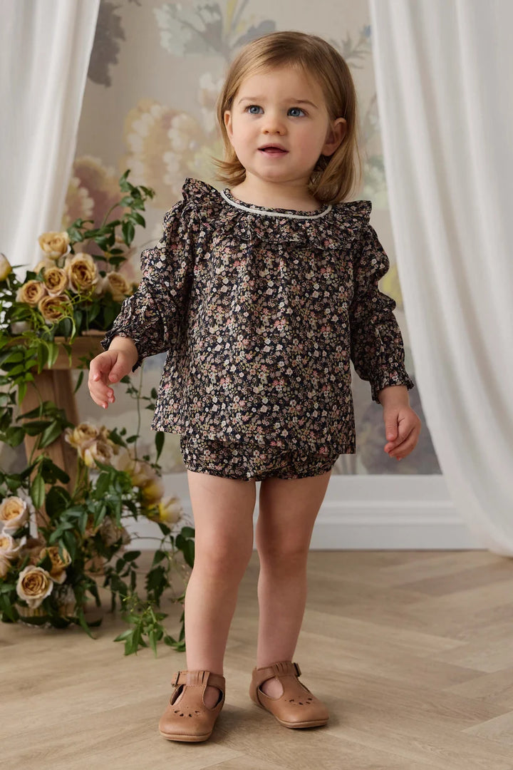 A toddler girl wearing a dark brown floral print romper with ruffled shoulders and tan leather Mary Jane shoes, standing in a decorated room with a floral backdrop and curtains.