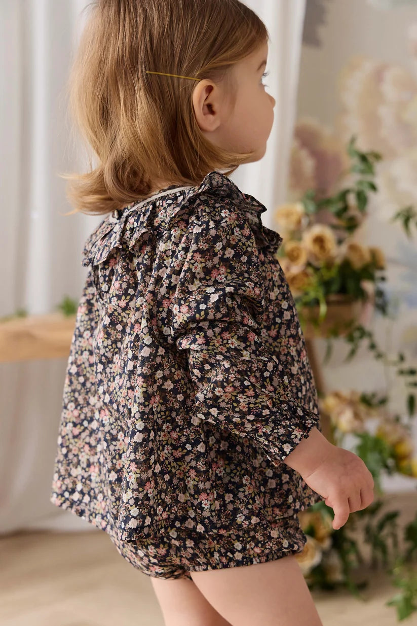 A young child wearing a dark floral print long-sleeve romper, photographed from the side in a softly lit indoor setting with flowers and curtains in the background.