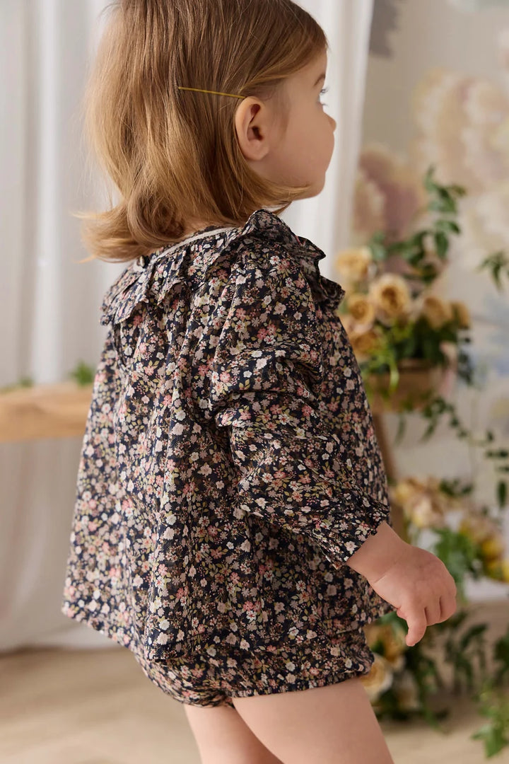 A young child wearing a dark floral print long-sleeve romper, photographed from the side in a softly lit indoor setting with flowers and curtains in the background.